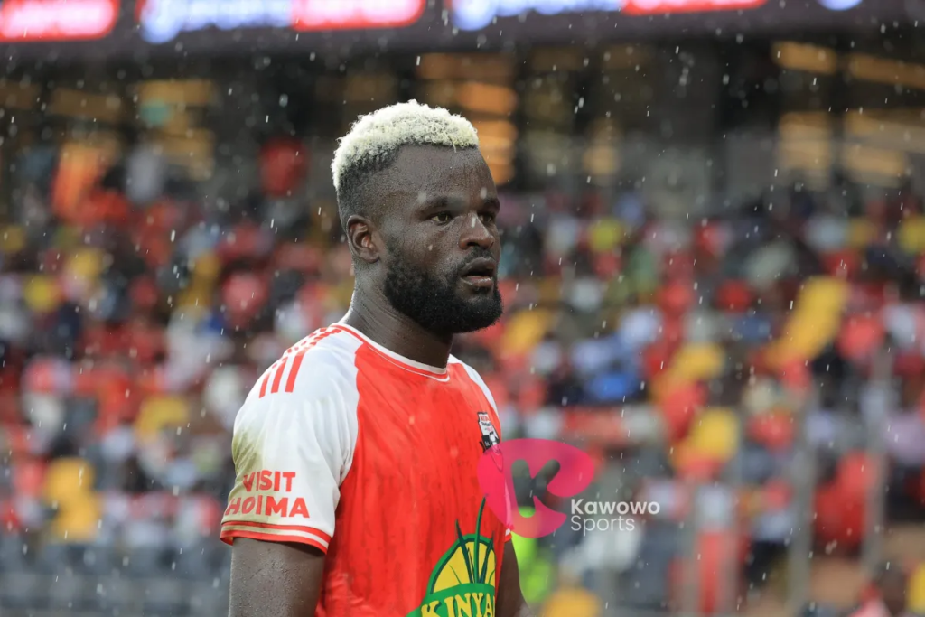 Gadafi Wahab, a bearded player with bleached blond hair, in a red and white Kitara kit with “Visit Hoima” on the sleeve, looking to the side as rain falls in front of a blurred crowd.