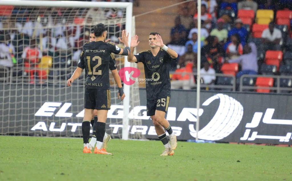 Two Al Ahly players in black kits, No. 12 on the left and No. 25 on the right, high-fiving on the pitch in light rain, with a third teammate partly hidden behind No. 12 and spectators in red, yellow and dark seats behind them.