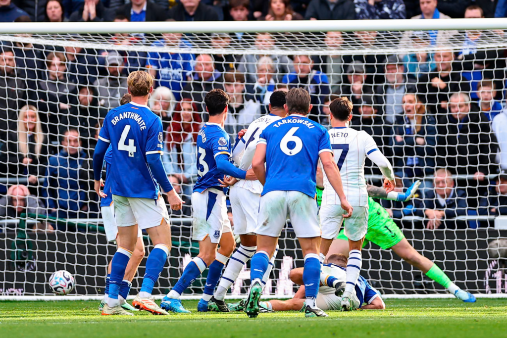 Seen from behind the goal net, the ball rests in the net at the bottom left as a goalkeeper in green lies stretched out on the ground to the right. Everton players in blue, including Branthwaite (No. 4) and Tarkowski (No. 6), stand watching, with Chelsea’s Neto (No. 7) in white among them. Another player lies on the ground and the stand behind is packed.