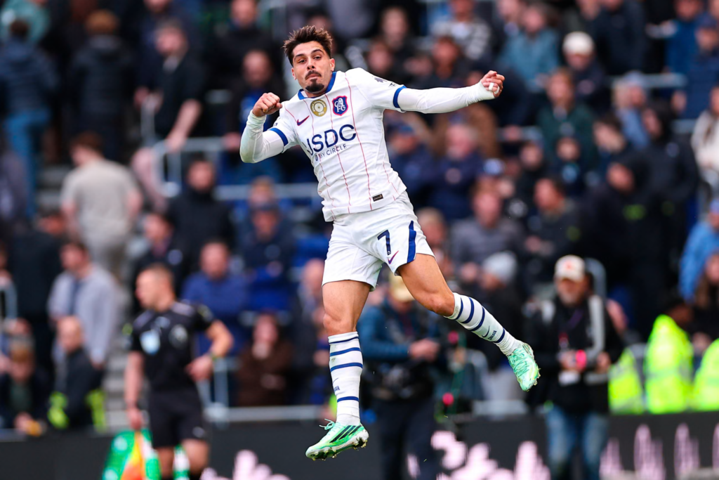 Chelsea’s Pedro Neto, wearing No. 7 in a white away kit, leaping in the air with both fists clenched and his mouth set, in front of a blurred crowd, a referee and photographers in high-vis jackets.