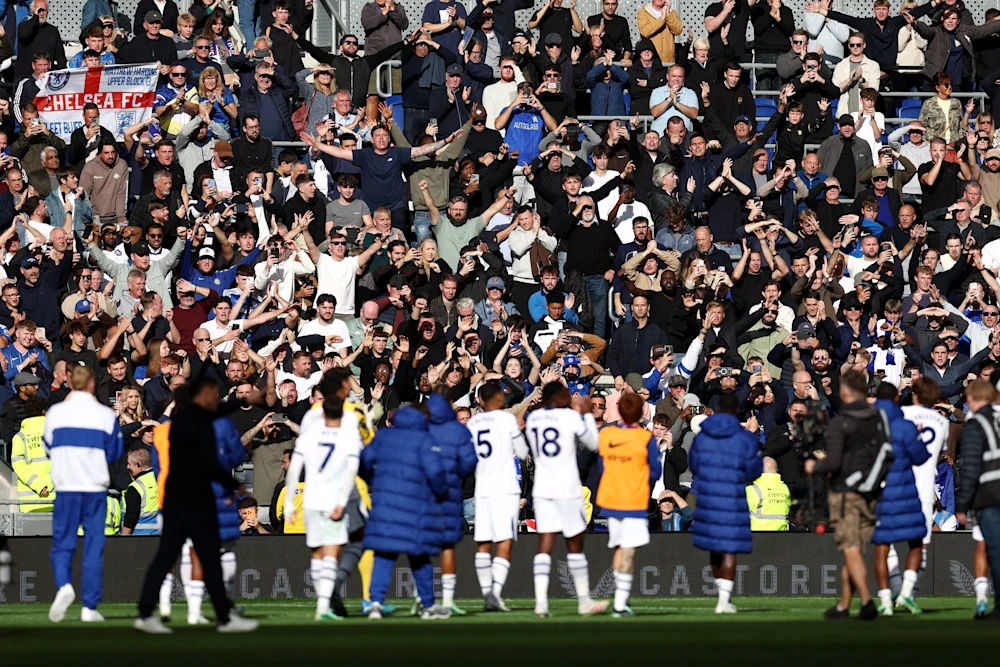 Chelsea players in white, including Nos. 7, 5 and 18, with staff in blue coats and a photographer in front of them, applauding the travelling fans at full-time. The packed stand is filled with supporters clapping and raising phones, with a “Chelsea FC” flag at the top left.