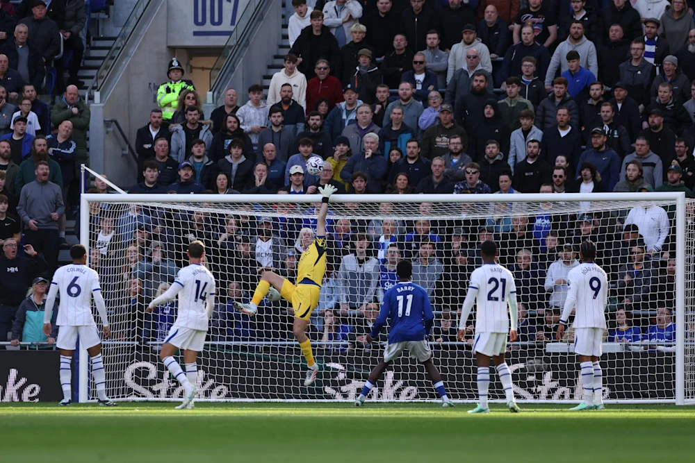 Chelsea goalkeeper Martinez, in yellow, leaping with one arm stretched high to reach the ball near the top of the goal. Chelsea’s Colwill (No. 6), a teammate in No. 14, Hato (No. 21) and João Pedro (No. 9), in white, stand around him, with Everton’s Barry (No. 11), in blue, between them and a packed stand behind the net.