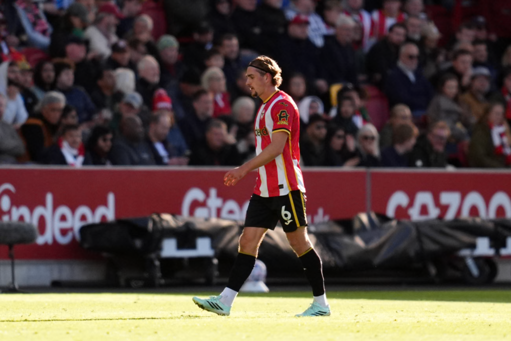 Brentford midfielder Yehor Yarmolyuk, wearing No. 6 in a red and white striped kit and a black headband, walking off the pitch with his head down in front of a seated crowd and red advertising boards