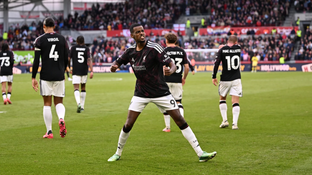 Liverpool’s Alexander Isak, wearing No. 9 in a dark kit, standing with his legs apart and fists clenched, teeth bared in celebration. Teammates with Virgil (No. 4), Jacquet (No. 5), Víctor M.V. (No. 23) and Mac Allister (No. 10) on their shirts walk away behind him, with a packed stand beyond them.