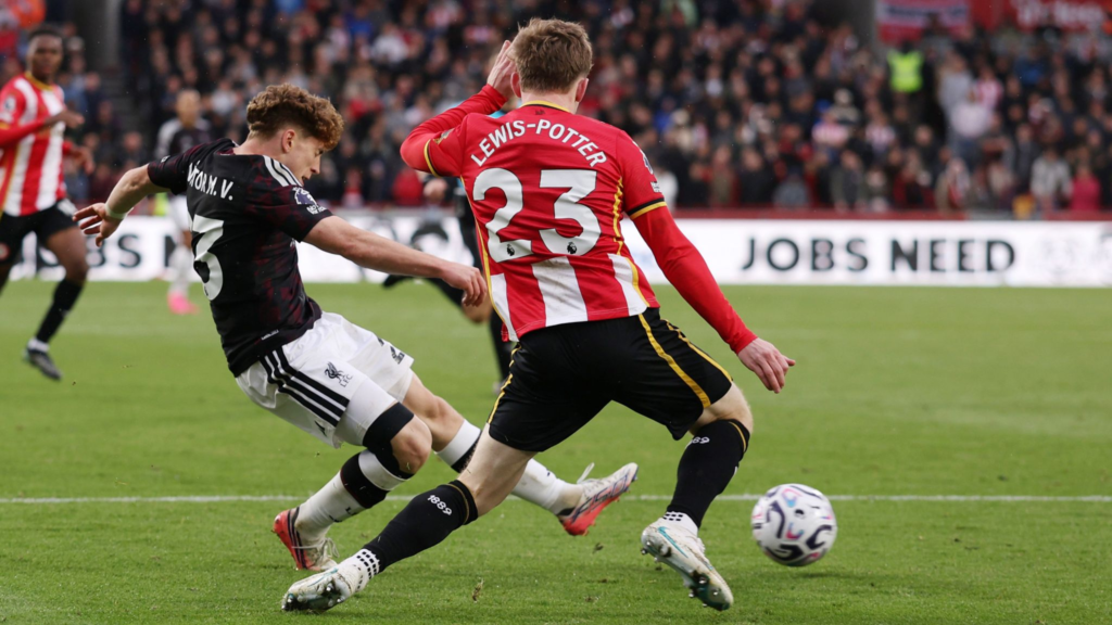 Liverpool’s Victor Munoz, in a dark kit with “Victor M. V.” on the shirt, leans back as he shoots. Brentford’s Lewis-Potter, in a red and white striped No. 23 shirt, stands in front of him with a leg stretched out. The ball is on the grass to the right, with a packed stand and an advertising board behind them.