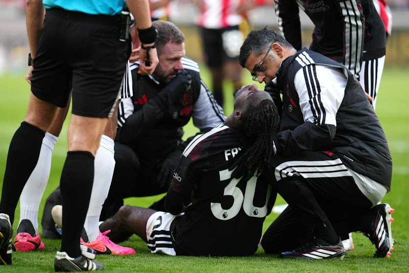 Liverpool’s Jeremie Frimpong, wearing No. 30 in a dark kit, sits on the grass with his head tilted back while two medical staff in black kneel beside him, one leaning in close. A referee in black shorts and several other players stand around them.