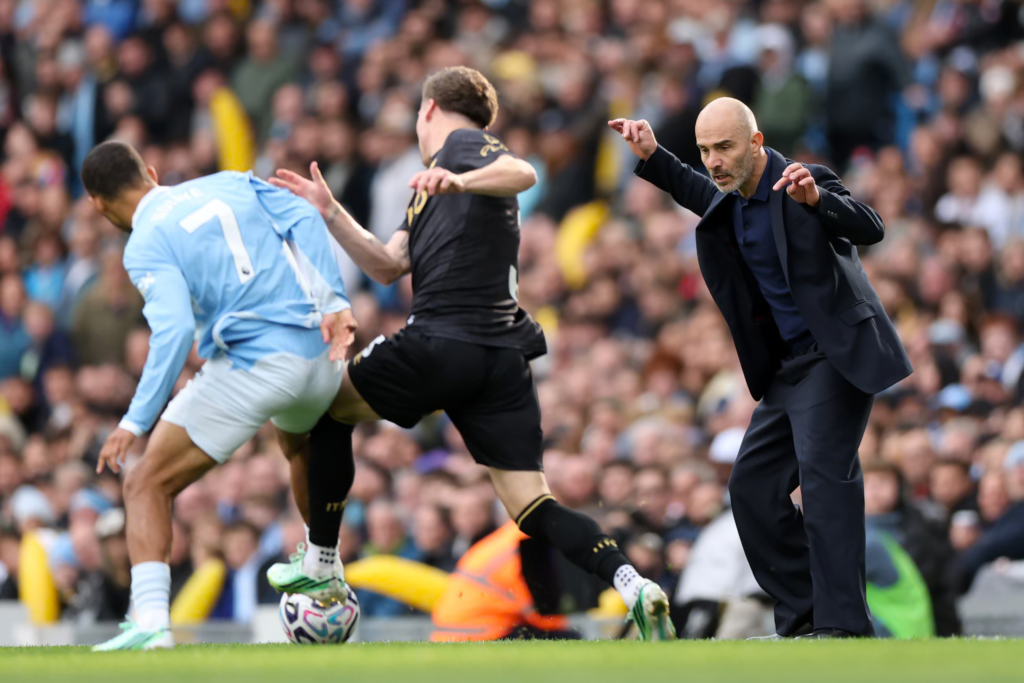Manchester City manager Enzo Maresca, in a dark navy suit, crouches on the touchline with both hands raised and his eyes on the pitch. Beside him, a City player wearing No. 7 in a sky blue kit and an opponent in black both go for the ball, which the opponent’s boot is on. A packed crowd fills the stand behind them.