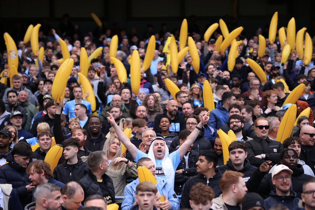 A packed stand of Manchester City fans, many in sky blue and dark jackets, hold up large yellow inflatable bananas. In the centre, a man in a sky blue shirt and a white headdress raises both arms with his index fingers pointing and his mouth open, shouting. Fans around him look on or smile.