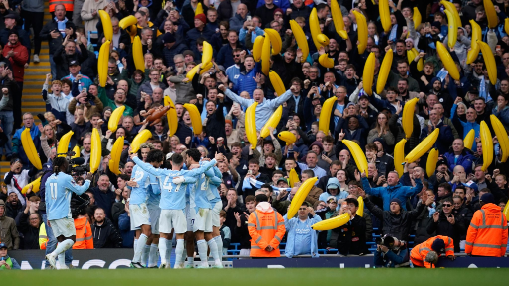 Manchester City players in sky blue kits, including Foden (No. 47), huddle with their arms around each other in front of a stand packed with cheering fans holding up large yellow inflatable bananas. To the left, a player wearing No. 11 with “Doku” printed on his shirt runs past a TV cameraman. Stewards in orange high-visibility jackets and photographers stand along the front of the stand.