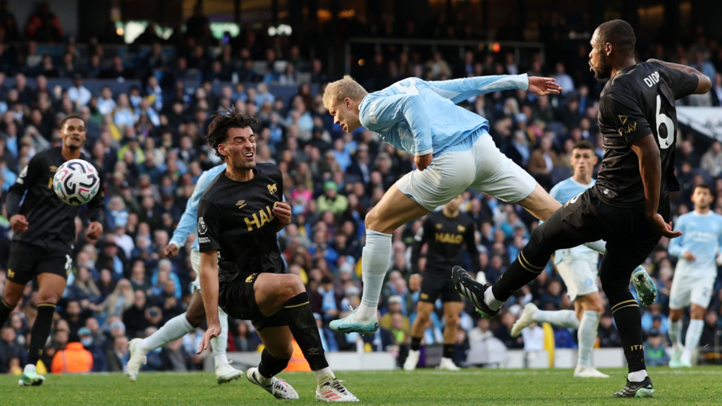 Manchester City’s Erling Haaland, in a sky blue kit, leaps and twists in mid-air with his arms out, his head turned toward the ball. Ipswich defender Diop, wearing No. 6 in black, sticks out a leg beside him. Another Ipswich player grimaces as he crouches back, and a ball is in the air at the left. Fans fill the stand behind them.