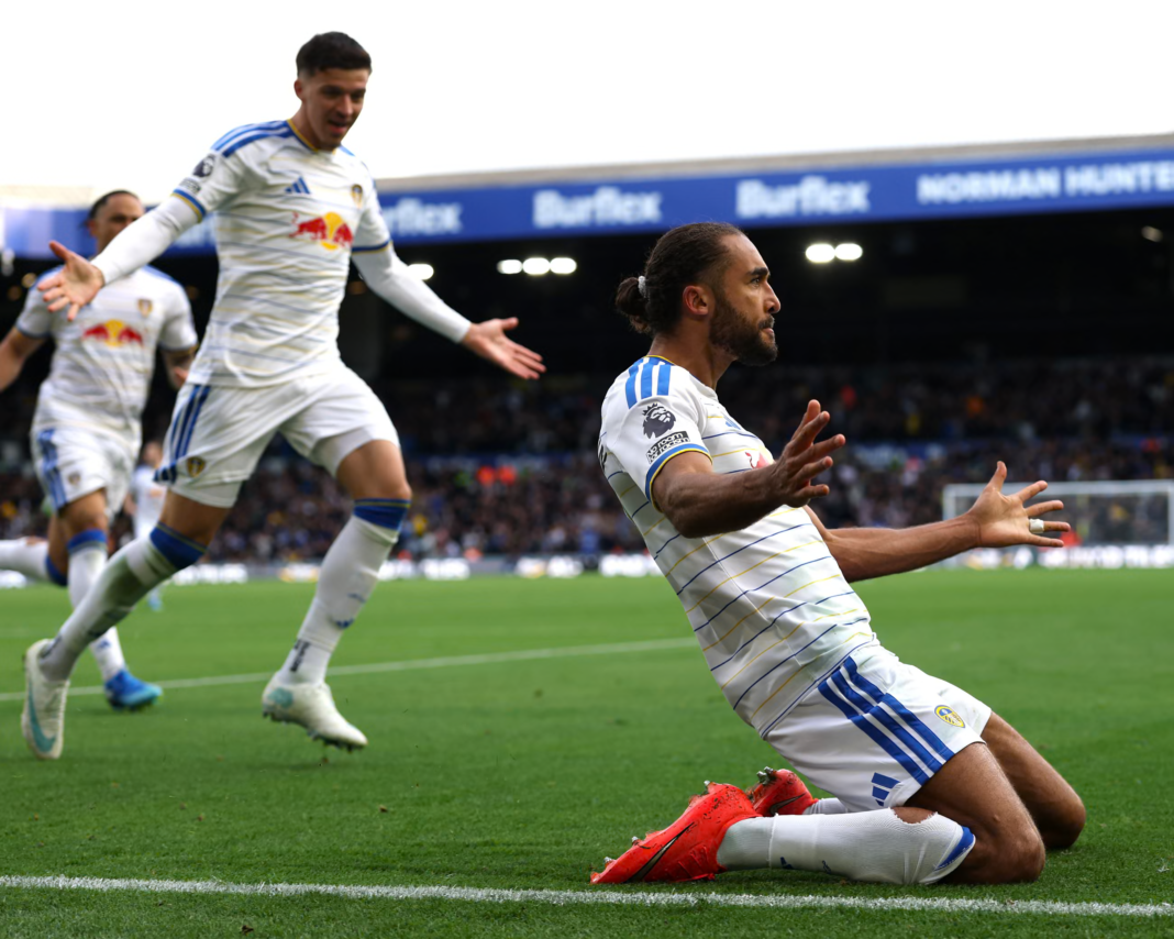 Leeds United’s Dominic Calvert-Lewin, in a white kit with blue stripes, kneels on the grass with both arms spread wide, his hair tied back and his eyes fixed ahead. Two teammates in the same kit run toward him behind, one with his arms open, with an empty goal and a stand in the background.
