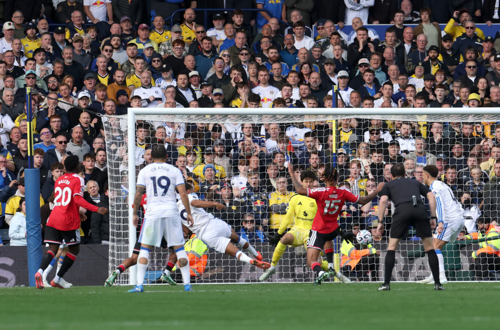 Leeds United’s Dominic Calvert-Lewin, in a white kit, stretches out almost horizontally in front of the goal as the ball reaches the foot of the net. Defenders in red shirts, Yoro (No. 15) and Baleba (No. 20), and a goalkeeper in yellow are around him. Leeds teammate Okafor (No. 19) stands in front, with a referee in black on the right. Behind the net, a packed stand of fans looks on, many with their hands raised to their faces.