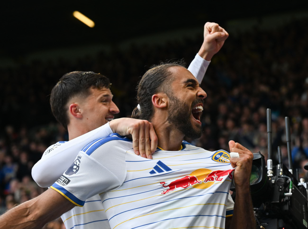 Leeds United’s Dominic Calvert-Lewin, in a white shirt with blue and yellow stripes, shouts in celebration with his mouth wide open, pulling the front of his shirt out with one hand. A teammate in the same kit stands behind him with an arm around his shoulder and a fist raised, smiling. A TV camera is at the right edge, with a dark crowd behind them.