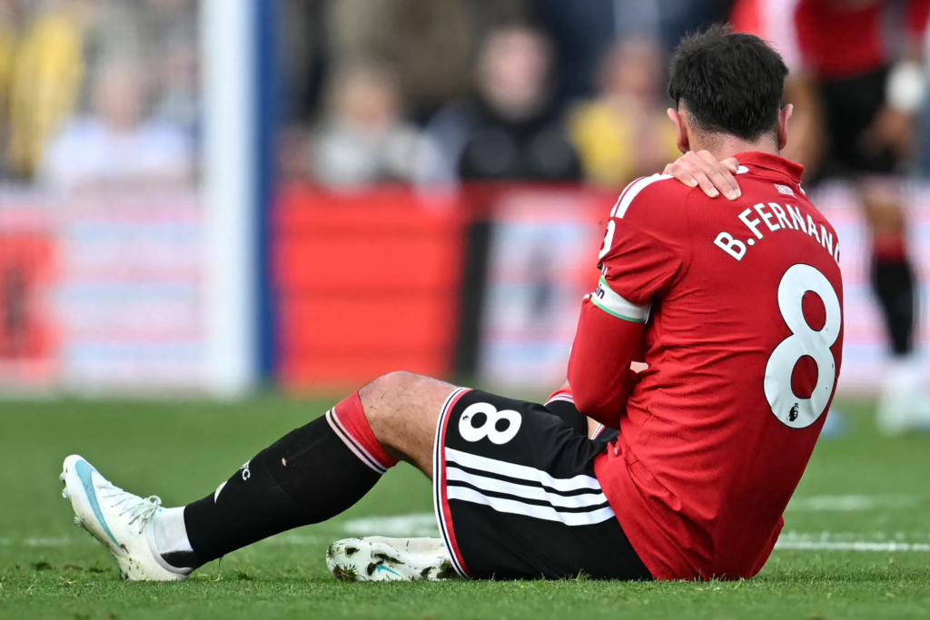 A player in a red kit with “B. Fernandes” and the number 8 on the back sits on the grass with his legs out in front of him, facing away from the camera. He has a captain’s armband on his left arm and holds his shoulder with one hand. The background of the pitch, advertising boards and crowd is blurred.