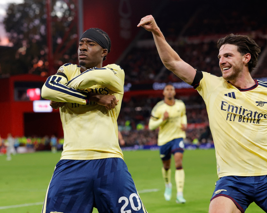 Arsenal’s Noni Madueke, in a pale yellow away shirt and navy shorts with No. 20, wears a black headband and crosses his arms over his chest with a serious expression. Teammate Declan Rice, in the same kit, leans in from the right with a raised fist and a broad smile. Another Arsenal player runs toward them in the background, with a blurred stand behind.