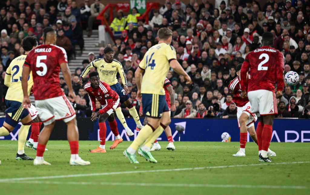 Arsenal’s Noni Madueke, in a pale yellow kit with a black headband, follows through with his right leg after striking the ball as defenders in red crowd around him. Foreground players include Arsenal’s No. 14 and No. 23 and red-shirted Murillo (No. 5) and O. Diomande (No. 2). A ball is in the air at the right edge, with a packed stand behind.