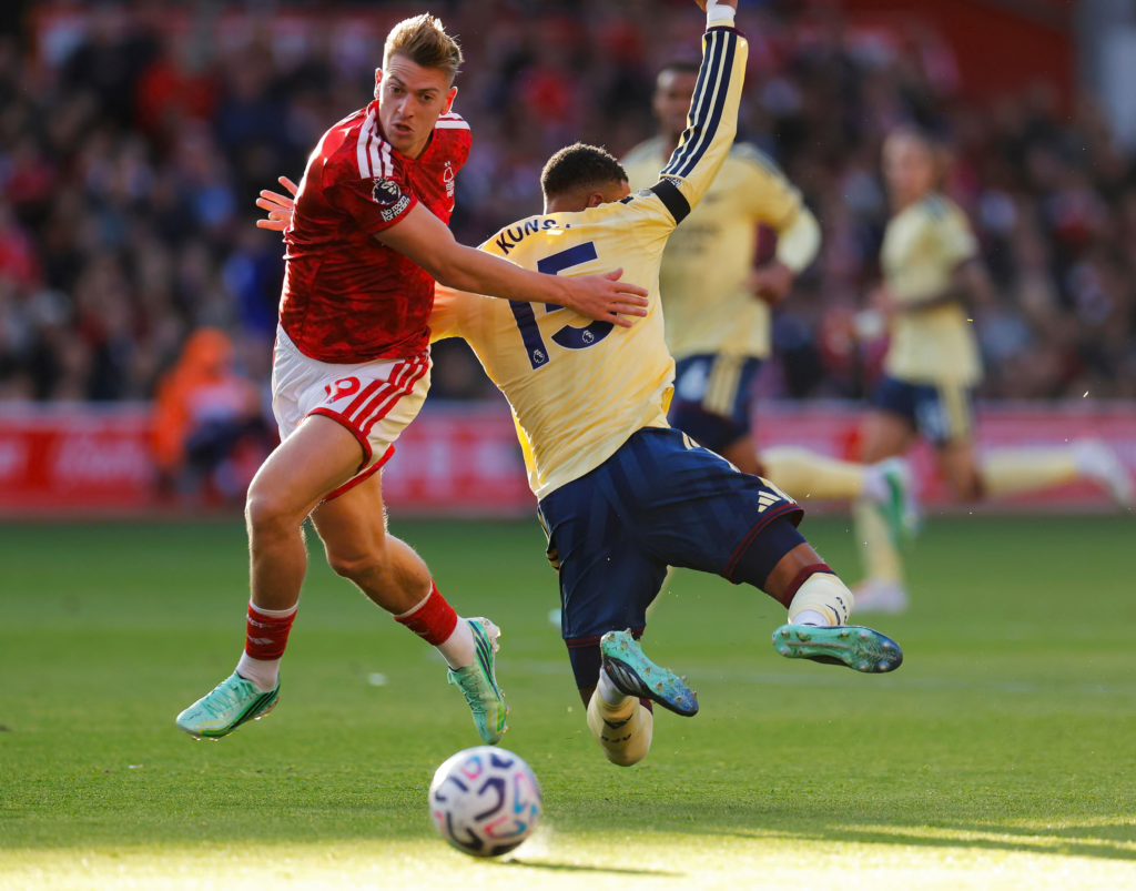 Liam Delap, in a red shirt and white shorts, strides forward with an arm out as Ezri Konsa, in a pale yellow shirt with “Kons…” and the number 15 on the back and navy shorts, tips backwards with a raised arm and both boots off the ground. A ball is on the grass in front of them, with a blurred teammate in yellow and a crowd behind.
