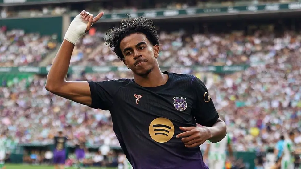 Barcelona’s Lamine Yamal, in a black and purple kit with the Spotify logo on the chest, points a finger up beside his head with a taped hand and looks ahead with a set expression. A packed stadium is blurred behind him, with teammates and opponents in green and white on the pitch.