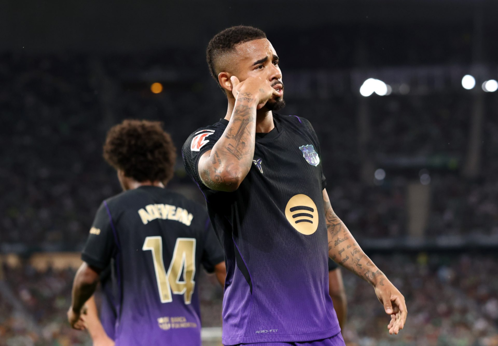 Barcelona’s Gabriel Jesus, in a black and purple kit with the Spotify logo on the chest, holds a hand with a tattooed forearm to his ear, looking to the side. A teammate wearing No. 14 stands with his back to the camera in the blurred foreground, with a dark stadium and floodlights behind.