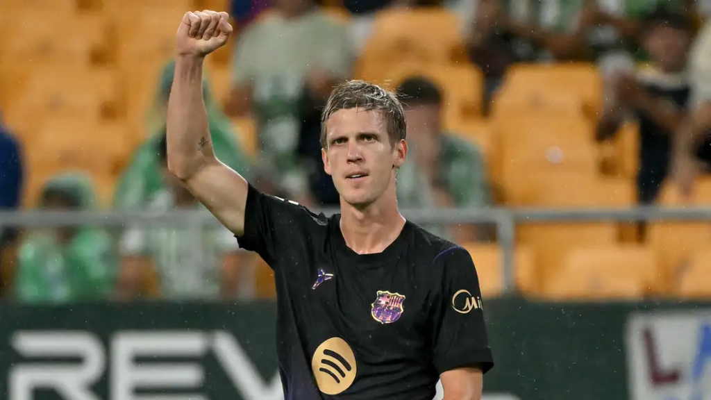 Barcelona’s Dani Olmo, in a black kit with the Spotify logo on the chest and wet hair, raises a clenched fist beside his head and looks ahead with a serious expression. Empty orange seats and a few fans in green and white are blurred behind him.