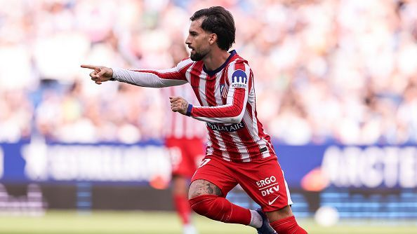 Atlético Madrid’s Alex Baena, in a red and white striped shirt and red shorts, runs with one arm pointing out to the side and the other bent at his chest, looking ahead with a beard and dark swept-back hair. A teammate in the same kit is partly visible behind him, with a blurred crowd and advertising boards in the background.