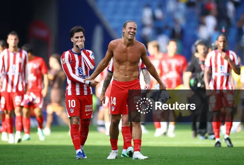 Atlético Madrid’s Alex Baena, wearing No. 10 in a red and white striped shirt, covers his mouth with a hand and smiles as Marcos Llorente, shirtless and in red shorts with No. 14, laughs beside him with his arms out. Teammates in red and white and a camera operator stand around them on the pitch, with blurred stands behind.