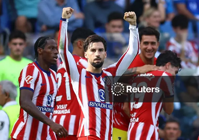 Atlético Madrid’s Alex Baena, in a red and white striped shirt with the “Riyadh Air” sponsor, raises both fists above his head with his eyes on the camera. Teammates in the same kit surround him, one on the left with braided hair, another behind him smiling and one on the right wearing No. 19. Fans fill the stand behind.