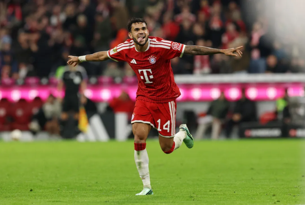 Bayern Munich’s Luis Díaz, in a red kit with the Telekom logo and No. 14 on his shorts, runs across the pitch with his arms stretched out wide and his tongue out, smiling. Fans and a pink-lit advertising board are blurred behind him.