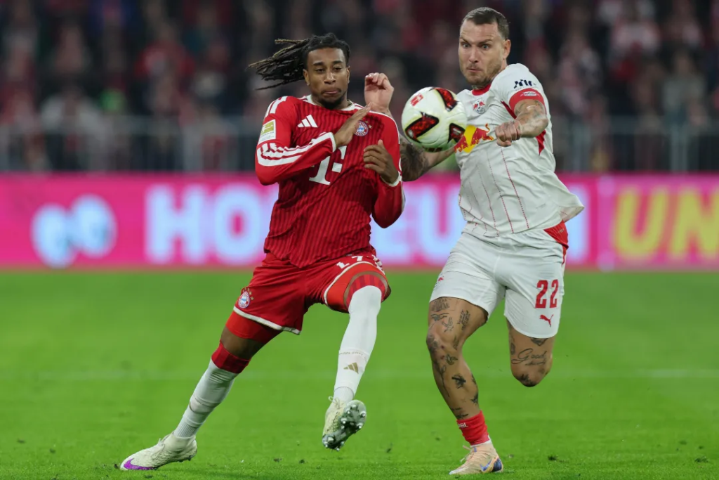 Bayern Munich’s Olise, in a red kit with his braids flying, and an RB Leipzig player in a white kit with No. 22 on his shorts both go for the ball in mid-air. The Leipzig player’s tattooed arm is raised, with a pink advertising board and blurred crowd behind them.