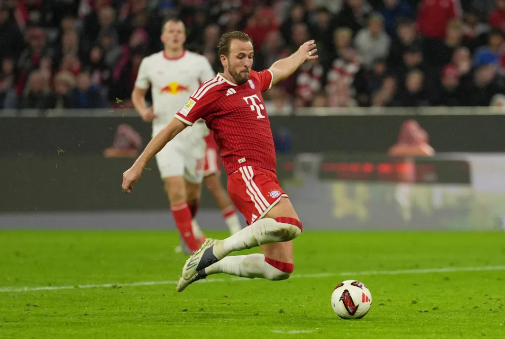 Bayern Munich’s Harry Kane, in a red kit with the Telekom logo, stretches out his right leg to strike a ball on the grass, with his left arm out for balance. An RB Leipzig player in a white kit with the Red Bull logo stands blurred behind him, with a crowd in the stand.