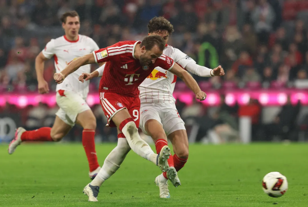 Bayern Munich’s Harry Kane, wearing No. 9 in a red kit with the Telekom logo, swings his right leg through as he shoots, with both arms out. An RB Leipzig player in white with the Red Bull logo is pressed against him, another Leipzig player runs behind, and the ball is on the grass at the right. A pink advertising board and crowd are blurred in the background