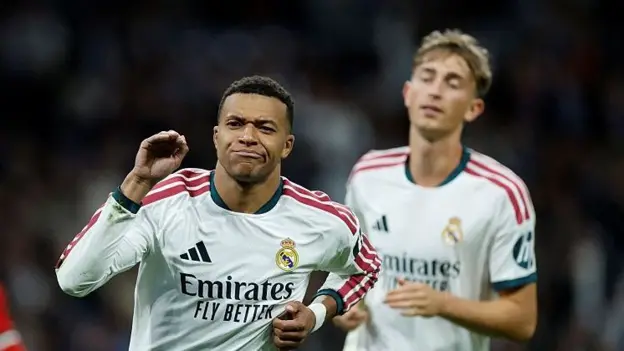 Real Madrid’s Mbappe, in a white kit with the Emirates logo, pumps a fist at his shoulder with his lips pressed together. A teammate in the same kit, with light brown hair, walks behind him, with a dark stadium blurred in the background.