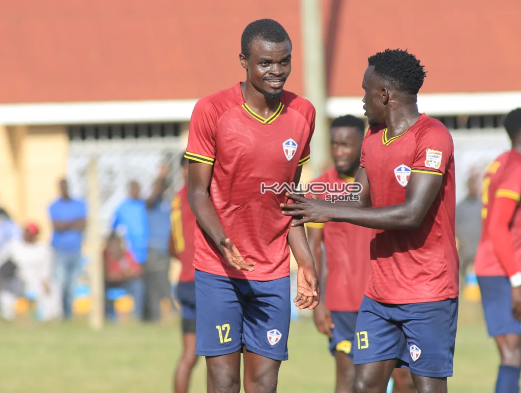 Two Maroons players in maroon shirts with yellow trim and navy shorts stand face to face. The player on the left, wearing No. 12, smiles with his open hands out. The player on the right, wearing No. 13, reaches a hand toward his chest. Teammates stand behind them, with a few spectators and a red-roofed building blurred in the background.