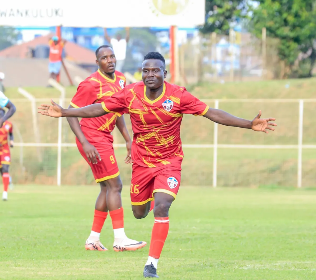 Fred Amaku, in a red shirt with gold streaks and red shorts marked No. 16, runs across the pitch with both arms stretched out wide. A teammate in the same kit, wearing No. 9, follows behind him. A fence, a grassy bank, a sign and trees are in the background.