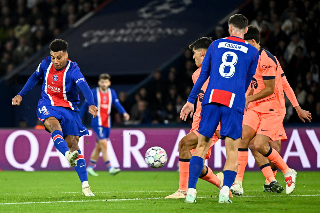 Paris Saint-Germain’s Désiré Doué, wearing No. 14 in a blue shirt with a red and white stripe, swings his right leg through to strike the ball on the grass. A PSG teammate with “Fabian” and No. 8 on his back stands with his back to the camera, with three players in orange kits crowding in front of him. A Qatar Airways advertising board and a Champions League banner are behind them.
