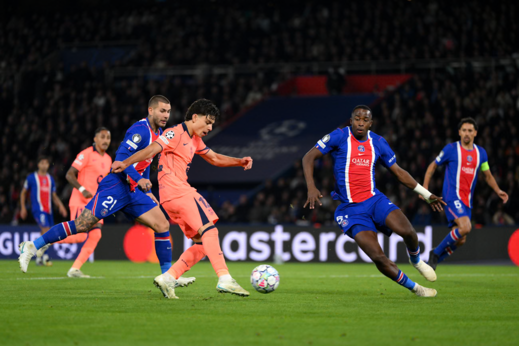 Barcelona’s Xavi Espart, in an orange kit with the Champions League badge on the sleeve, strikes the ball with his right foot as PSG defenders in blue shirts with a red and white stripe close in. One PSG player in No. 21 shorts is beside him, another in No. 51 shorts stretches a leg out on the right, and a captain with No. 5 runs in behind. A Mastercard advertising board and a dark, packed stand are in the background.