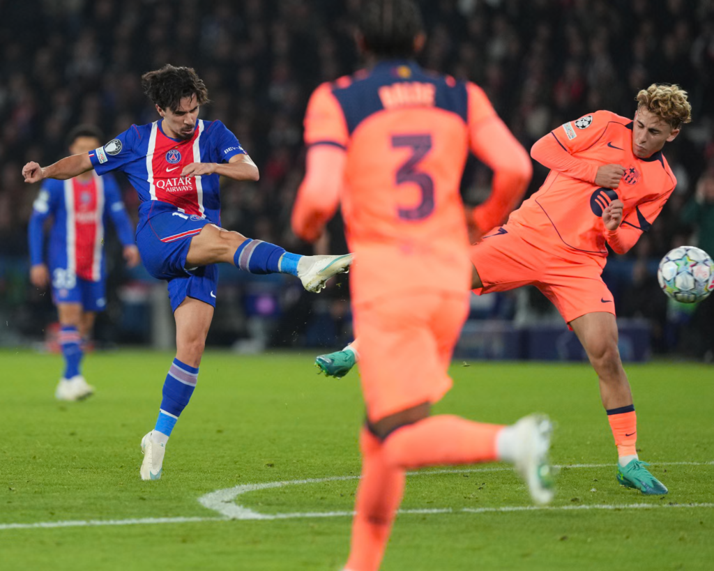 Paris Saint-Germain’s Vitinha, in a blue shirt with a red and white stripe, swings his right leg out to strike the ball with a captain’s armband on his left arm. Two Barcelona players in orange kits are in front of him, one blurred in the foreground with No. 3 on his back and another on the right with curly blond hair, leaning away as the ball flies past at the right edge. A PSG teammate is blurred behind, with a dark stadium behind them.