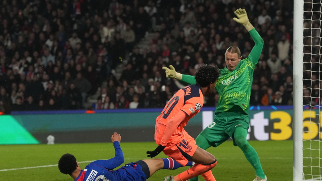 Barcelona’s Lamine Yamal, in an orange kit with “Yamal” and No. 10 on the back, moves in on goal as the PSG goalkeeper in a green kit with No. 29 throws both arms up. A PSG player in blue with “Zaire-Emery” partly visible on his shirt lies on the grass beneath them. The goalpost and net are at the right, with a packed stand behind.