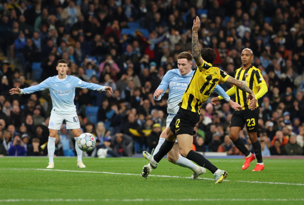 Manchester City’s Elliot Anderson, in a sky blue kit, strikes the ball with a fist clenched as an opponent in a yellow and black kit with No. 12 stretches across him with a tattooed arm raised. A City teammate wearing No. 47 stands on the left with his arms out, and another opponent wearing No. 23 watches from behind. A ball sits on the grass at the left, with a packed stand in the background.