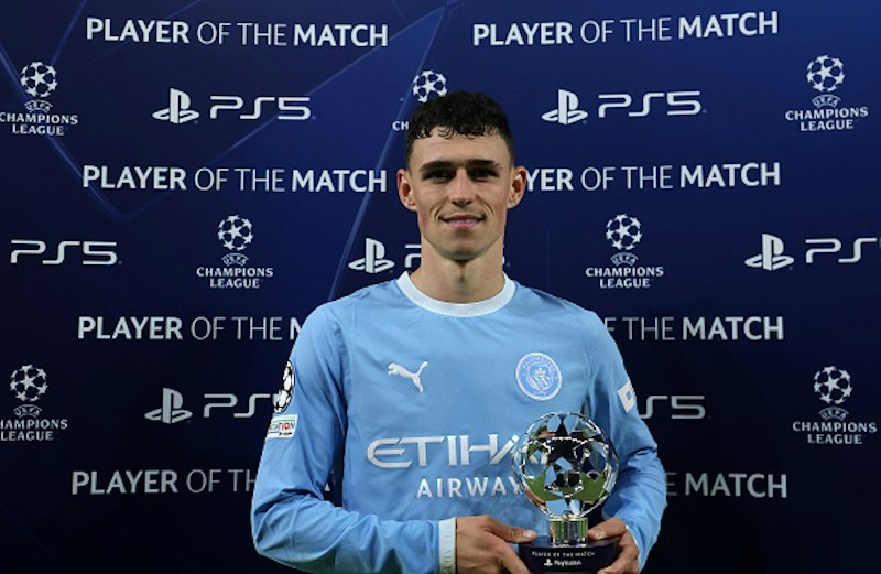 Manchester City’s Phil Foden, in a sky blue long-sleeved kit with the Etihad Airways logo, stands smiling and holds a small Champions League Player of the Match trophy in front of him. Behind him is a dark blue backdrop repeating the words “Player of the Match” with the Champions League and PS5 logos.