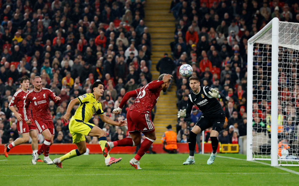 Liverpool’s Ronald Araujo, in a red kit with a bandaged wrist, leans back as he heads the ball in front of the goal, with a Villarreal goalkeeper in black reaching toward it at the right. A Villarreal player in yellow runs beside him, two Liverpool teammates in red follow from the left, and a packed Anfield stand fills the background.