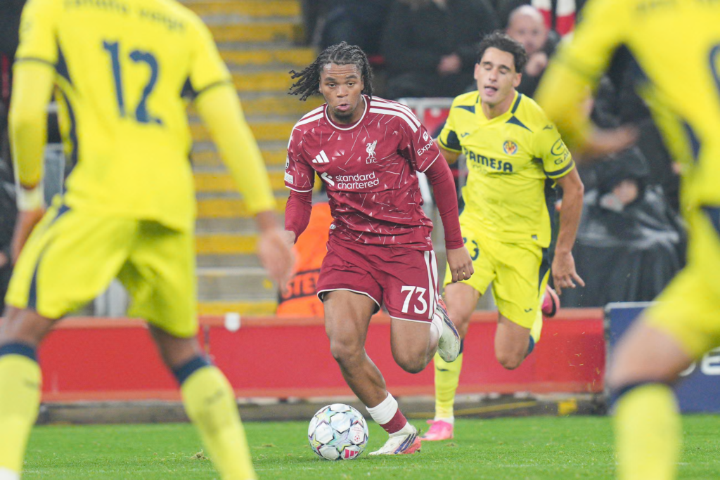 Liverpool’s Rio Ngumoha, wearing No. 73 in a red patterned kit with a taped sock, dribbles the ball toward the camera with his braids flying behind him. Villarreal players in yellow surround him, two blurred in the foreground, one with No. 12 on his back, and another chasing from behind on the right. An Anfield stand and red advertising boards are in the background.