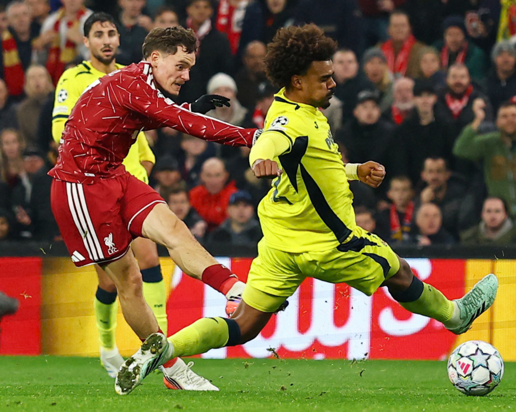 Liverpool’s Florian Wirtz, in a red kit, swings his right leg through as he shoots, his eyes on the ball. A Villarreal player in a yellow kit with curly hair slides across him with a leg stretched out, and another player in yellow watches from behind. The ball is on the grass at the bottom right, with fans in the stand and red advertising boards behind.