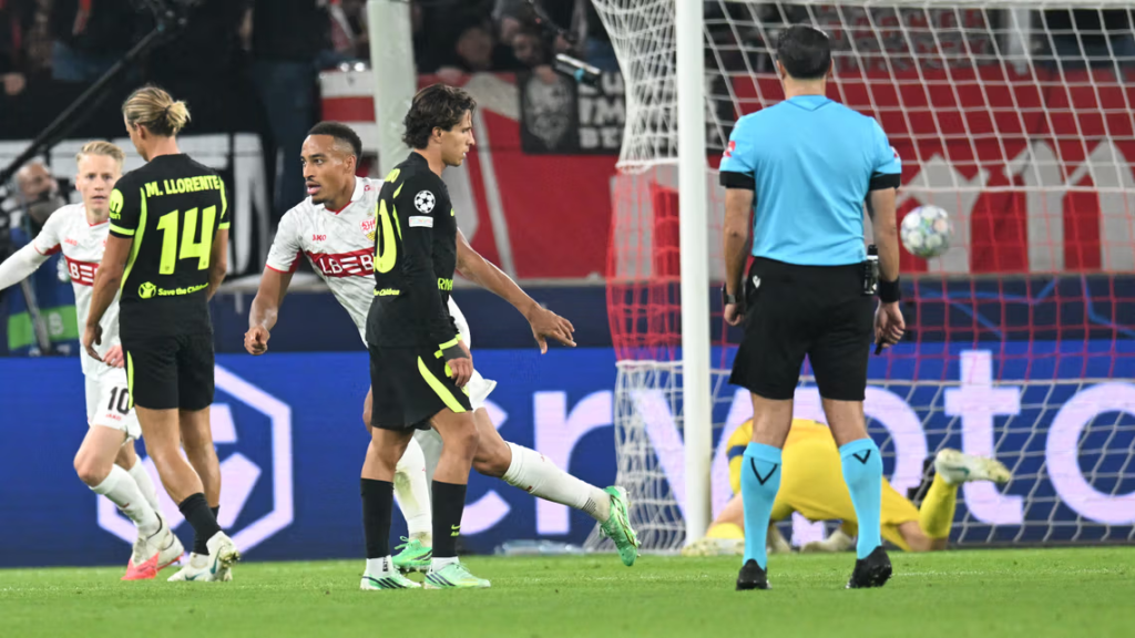 Atlético Madrid players in black and neon green kits, including M. Llorente with No. 14 and another with No. 10, stand and walk on the pitch near the goal. Stuttgart players in white shirts with a red chest stripe are close beside them, one with his arm out. A goalkeeper in yellow lies on the grass in the goal, a referee in light blue stands with his back to the camera, and a ball is in the air by the post.