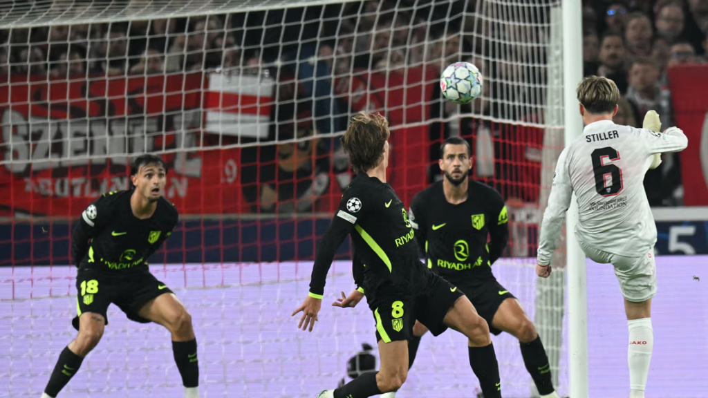 VfB Stuttgart’s Angelo Stiller, wearing No. 6 in a white kit with “Stiller” on the back, kicks the ball high with his right foot near the goalpost. Three Atlético Madrid players in black and neon green kits, wearing No. 18, No. 8 and one other, stand in front of him and watch. The ball is in the air above them, with the goal net and a red banner in the stand behind.