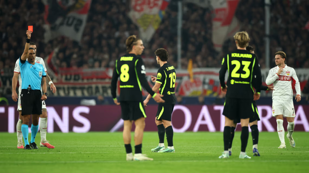 A referee in a light blue shirt holds up a red card in his raised right hand as he faces the camera, with a Stuttgart player in white beside him. Atlético Madrid players in black and neon green kits stand in a group, with No. 8 and No. 19 facing the camera and No. 23, “Hjulmand”, in the foreground with his back turned. Another Stuttgart player in white walks on the right, with a crowd and banners behind.