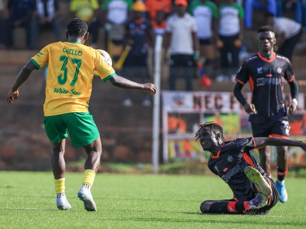 A BUL player in a yellow shirt with green trim, “Okello” and No. 37 on the back, walks away from the camera with his arm out, with the ball just beyond his shoulder. An NEC player in a black and orange kit slides along the grass on the right with one arm stretched out, and a teammate in the same kit stands behind him. A few spectators and an advertising board are in the background.