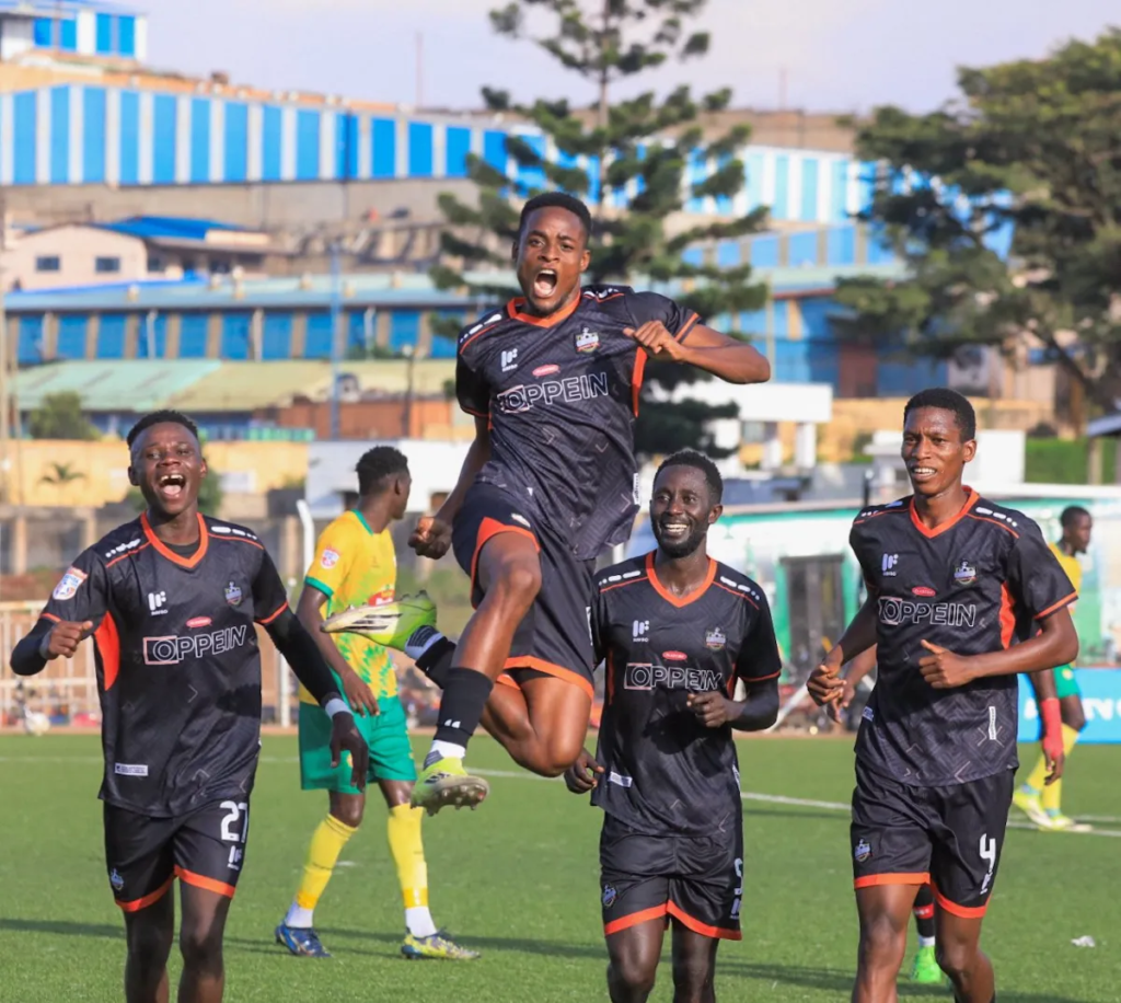 A player in a black and orange kit with “Oppein” on the chest leaps off the ground with a fist clenched and his mouth open in celebration. Three teammates in the same kit run around him, smiling, wearing No. 27 and No. 4. Two players in yellow and green kits are behind them, with a pine tree and blue-roofed buildings in the background.