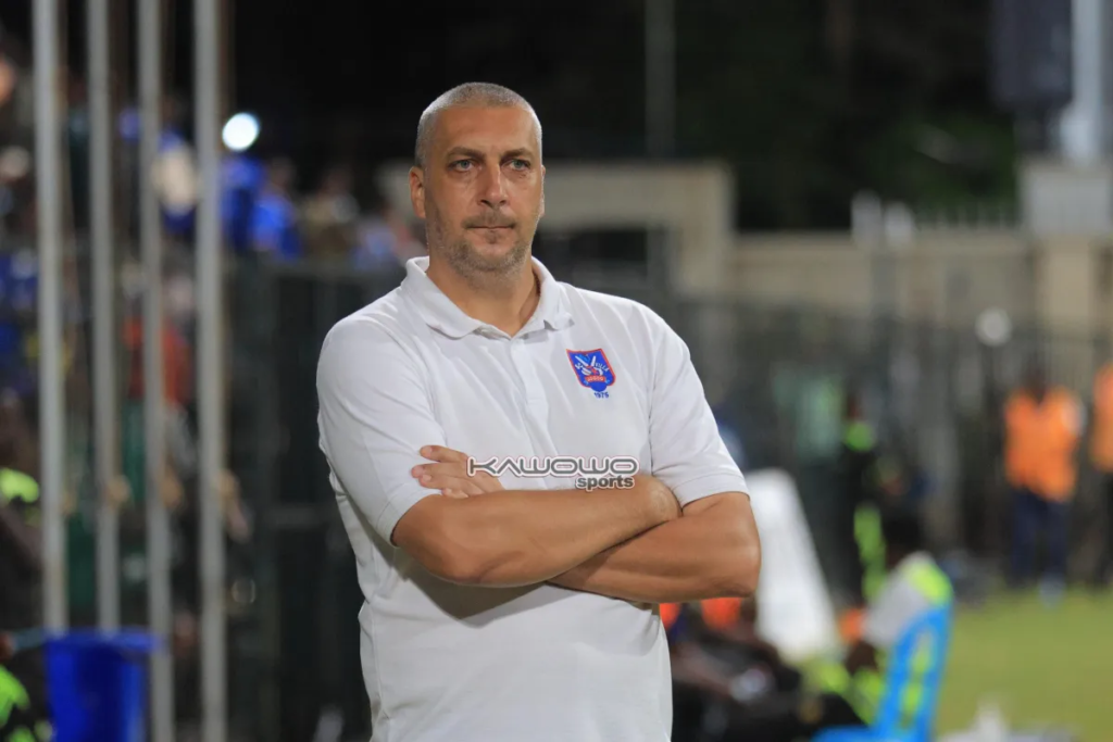 SC Villa coach Kovacevic Zeljko, a man with short grey hair and stubble, stands in a white polo shirt with a club crest on the chest and his arms folded, looking to the side with a serious expression. Blurred behind him are a fence, spectators in blue and staff in orange high-visibility vests, with a blue plastic chair at the bottom right.