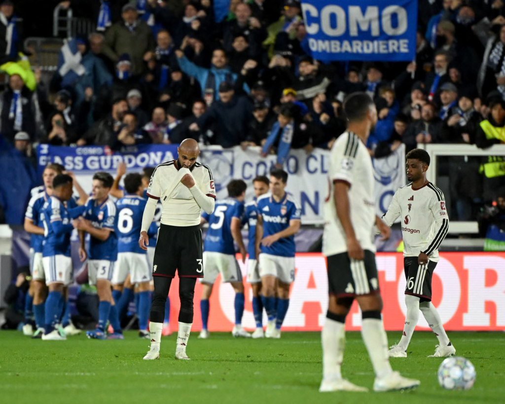 Three Manchester United players in white shirts and black shorts stand dejected on the pitch. The one in the centre, wearing No. 3, holds the neck of his shirt over his mouth with his head bowed. A second, in the foreground, is blurred, and a third wearing No. 16 stands on the right looking down. Behind them, Como players in blue and white huddle in celebration in front of a stand of fans holding blue banners, one reading “Como” and “Lombardiere”. A ball sits on the grass at the bottom right.